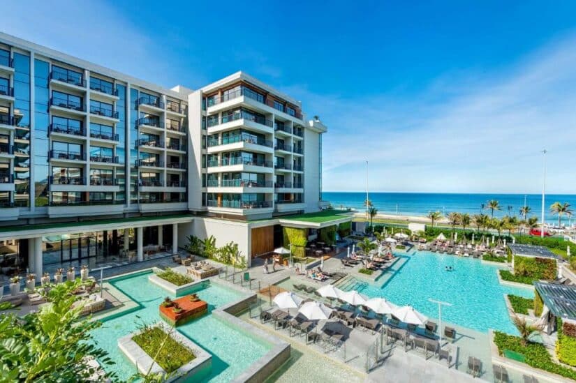 Hotel de luxo à beira-mar onde ficar na Barra da Tijuca com varandas modernas, piscinas externas, espreguiçadeiras e vista para o mar sob um céu azul claro.