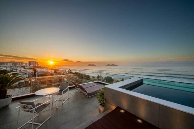 Pátio na cobertura com piscina e espreguiçadeiras onde ficar na Barra da Tijuca com vista para o oceano ao pôr do sol.