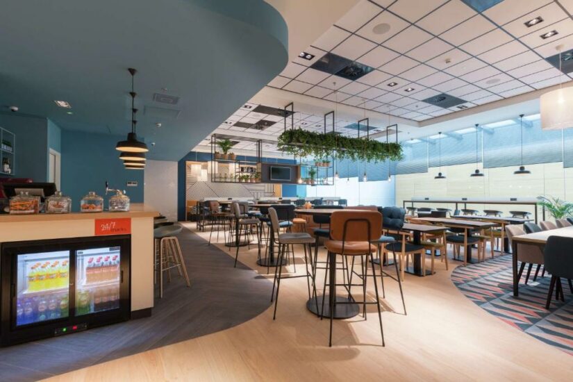 Lounge moderno e bem iluminado com mesas, cadeiras, frigobar e plantas suspensas em um ambiente espaçoso.