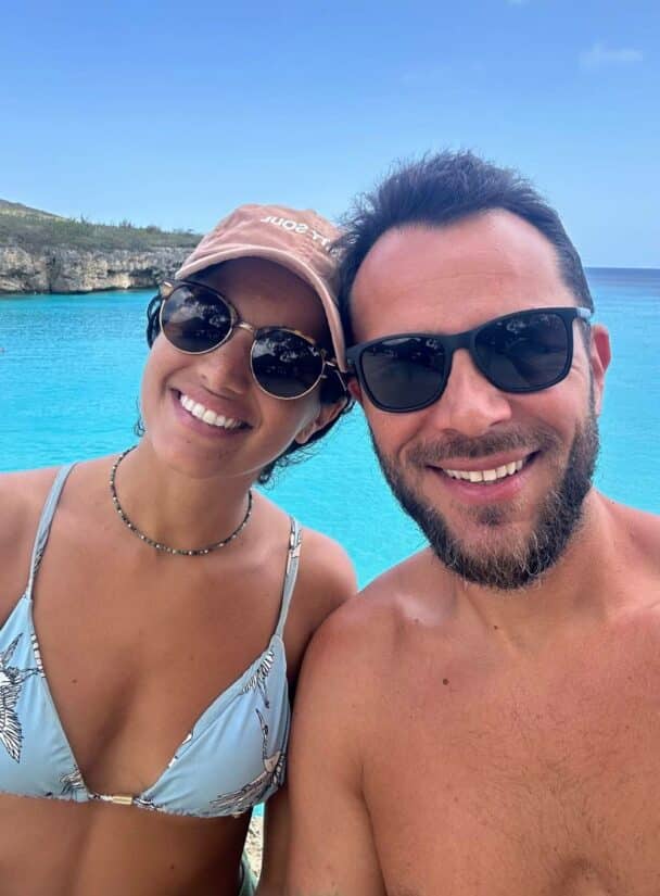 Um casal sorridente de óculos escuros posa em frente a uma água azul brilhante, sonhando com como funciona um Home Exchange, sob um céu claro.