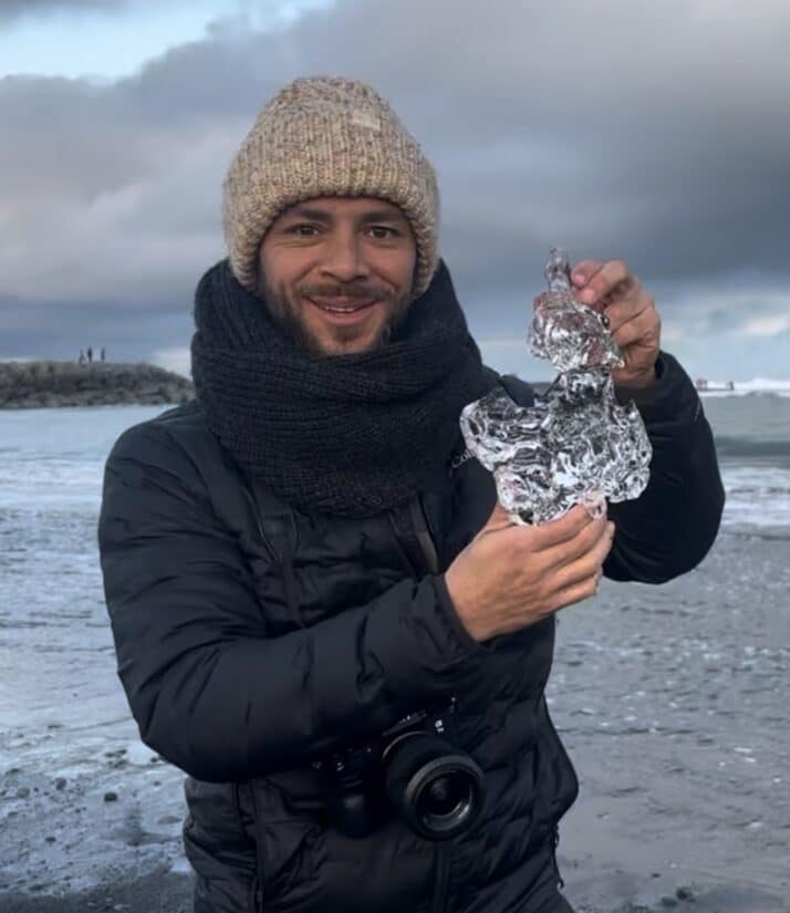 Homem sorridente em roupas de inverno segura um pedaço de gelo transparente em uma praia nevada, com uma câmera pendurada no pescoço.