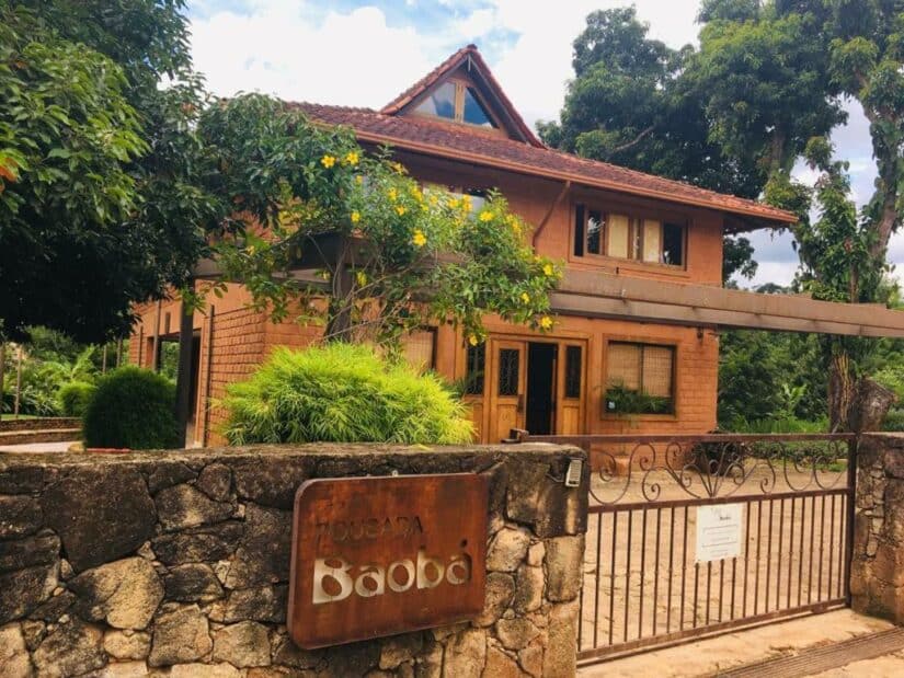 hotéis em Bichinho Minas Gerais Casa de dois andares com telhado de telha vermelha onde ficar em Bichinho, cerca de pedra e uma placa indicando Pousada Baobá na entrada.