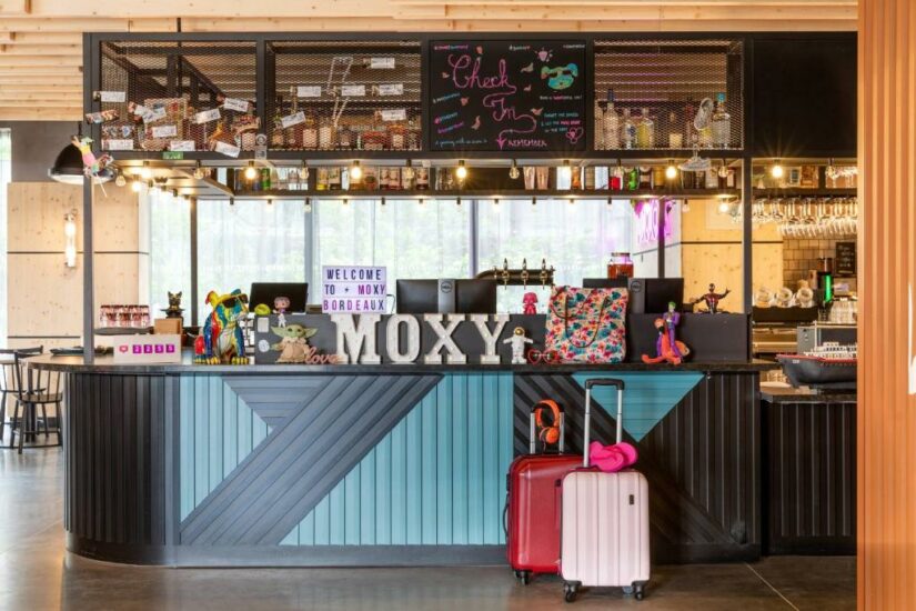 Recepção de hotel colorida onde ficar em Bordeaux com decoração divertida, placa MOXY e duas malas na frente.