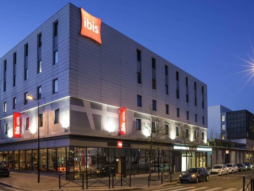 Moderno hotel Ibis ao anoitecer com letreiros iluminados, grandes janelas e carros estacionados na rua.