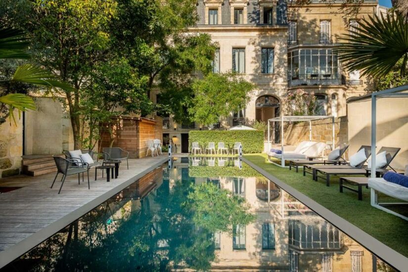 Elegante piscina externa com espreguiçadeiras, vegetação e um edifício histórico ao fundo em um dia ensolarado.
