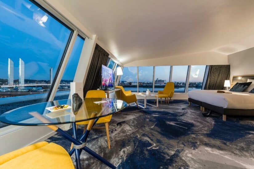 Quarto de hotel onde ficar em Bordeaux com janelas grandes, vista para a cidade, mesa redonda de vidro, cadeiras amarelas e uma cama com decoração moderna.