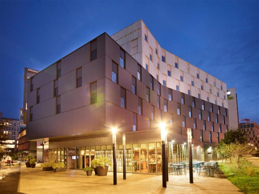 Hotel moderno ao entardecer, com área de estar ao ar livre — a opção ideal para se hospedar em Bordeaux, com entrada envidraçada e iluminação intensa.
