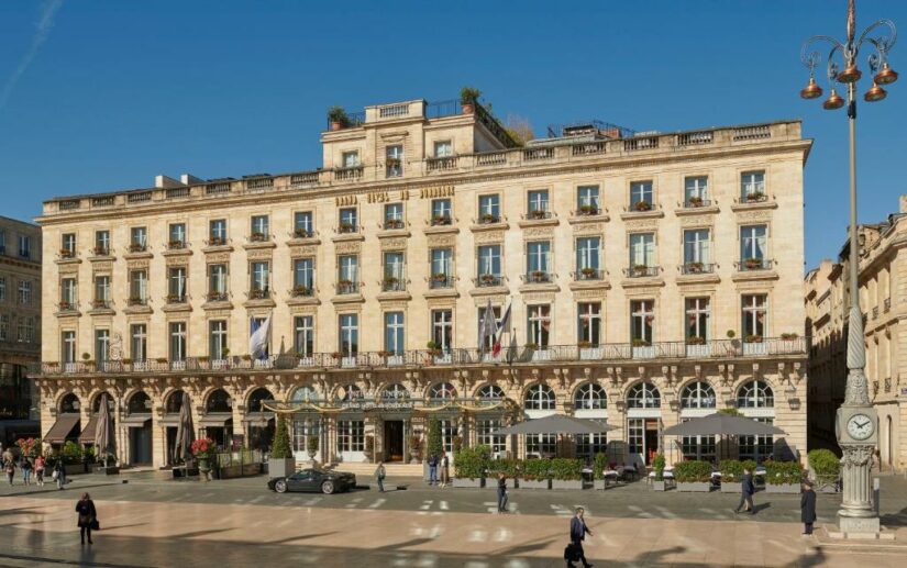 Edifício elegante e histórico do hotel onde ficar em Bordeaux, com janelas em arco e varandas, de frente para uma praça com pessoas e um relógio.