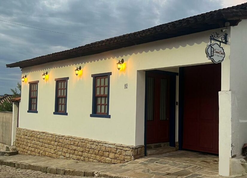 Edifício branco em estilo colonial onde ficar em Catas Altas com base de pedra, portas vermelhas, acabamento azul e luzes externas amarelas ao anoitecer.