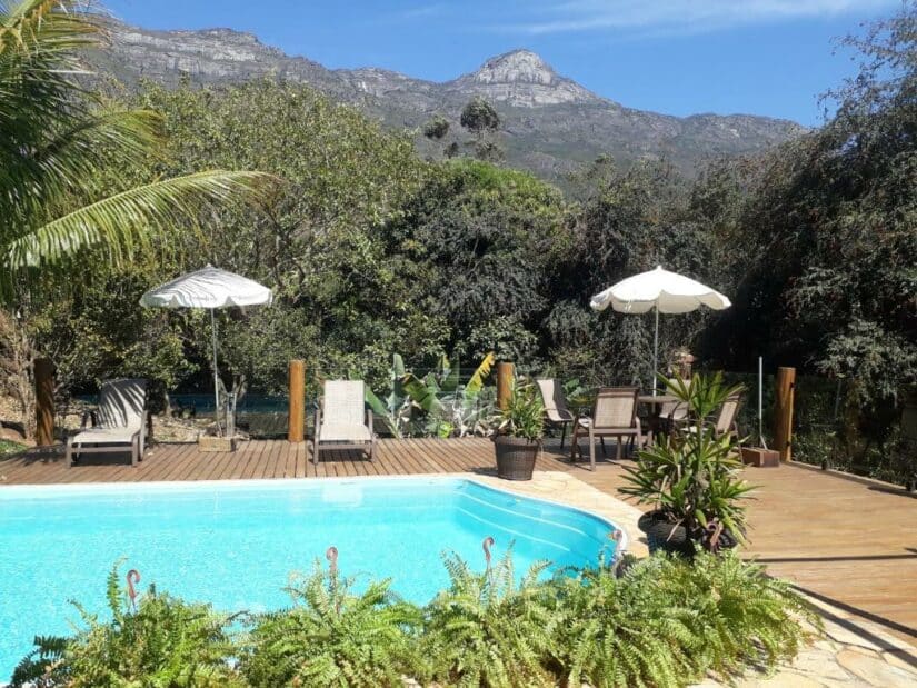 Uma piscina com espreguiçadeiras e guarda-sóis, cercada por vegetação e montanhas ao fundo.