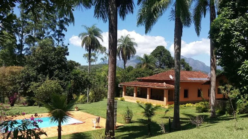 Uma casa onde ficar em Catas Altas com telhado de telha vermelha fica entre palmeiras perto de uma piscina, com montanhas ao fundo.