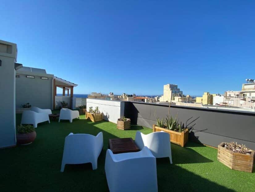 hotéis em Heraklião Grécia Terraço na cobertura com cadeiras brancas, vasos de plantas e vista para a cidade — um lugar ideal onde ficar em Creta.