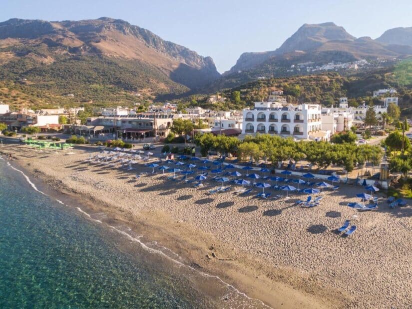 hotéis em Plakios na Grécia Praia de areia com fileiras de guarda-sóis azuis, águas cristalinas — perfeita para quem está se perguntando onde ficar em Creta em Plakias.