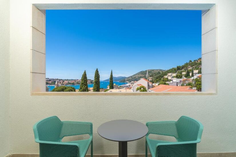onde ficar em Dubrovnik em Gruz Duas cadeiras turquesa e uma mesa em uma varanda em Gruz na Croácia têm vista para os telhados, árvores e o mar azul.