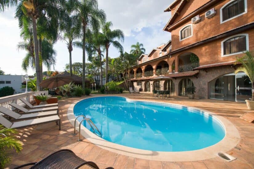 hotéis em São João da Boa Vista perto de Espírito Santo do Pinhal Piscina ensolarada em um resort onde ficar em Espírito Santo do Pinhal com espreguiçadeiras, palmeiras e um grande prédio de tijolos ao fundo.