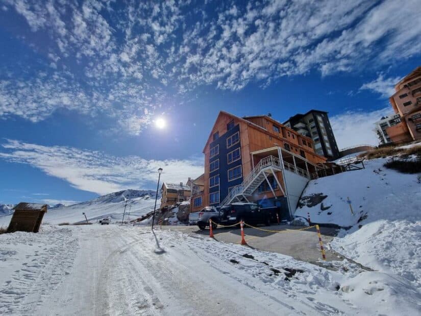 onde ficar em Farellones perto do Valle Nevado Uma estrada de montanha nevada com um prédio de madeira, carros estacionados e um sol brilhante em um céu azul com nuvens.