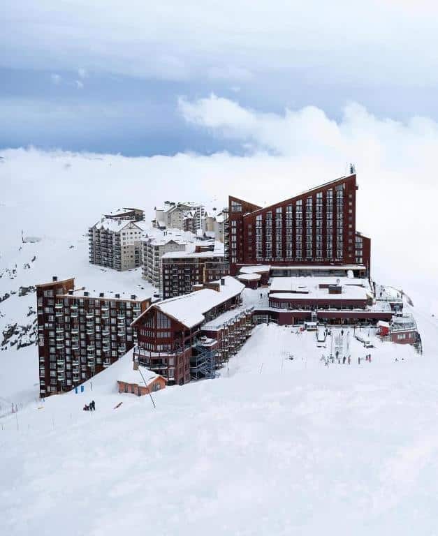 onde ficar em Farellones perto de Santiago no Chile Edifícios de estações de esqui cobertos de neve com um design moderno e angular, cercados por montanhas e céu nublado.