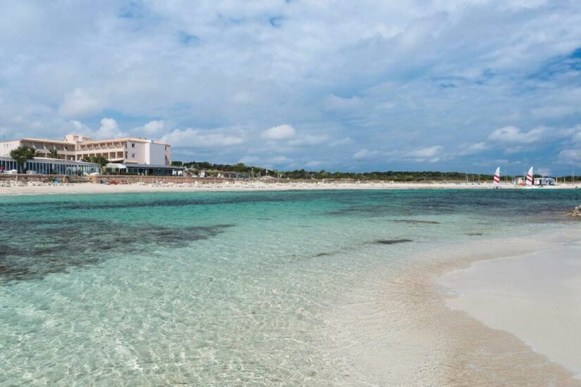 hotéis em Es Pujols em Formentera na Espanha Água azul-turquesa clara e praia de areia com um edifício de hotel onde ficar em Formentera e veleiros sob um céu parcialmente nublado.