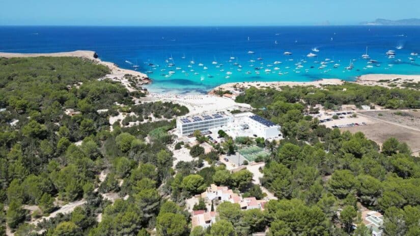 Vista aérea de onde ficar em Formentera: árvores verdes, uma praia de areia, mar azul-turquesa e muitos barcos ancorados.