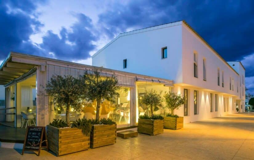 onde ficar em Formentera nas Ilhas Baleares Espanha Edifício branco moderno com janelas grandes, interior com iluminação aconchegante e vasos de plantas do lado de fora ao anoitecer.