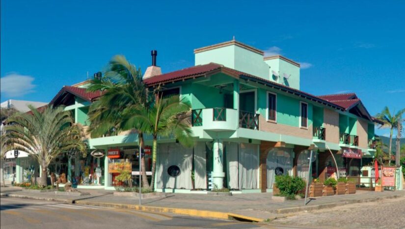 hotéis e pousadas no litoral de Santa Catarina Um prédio turquesa de dois andares com palmeiras, lojas e restaurantes - onde ficar em Garopaba na Praia Central em uma esquina ensolarada.