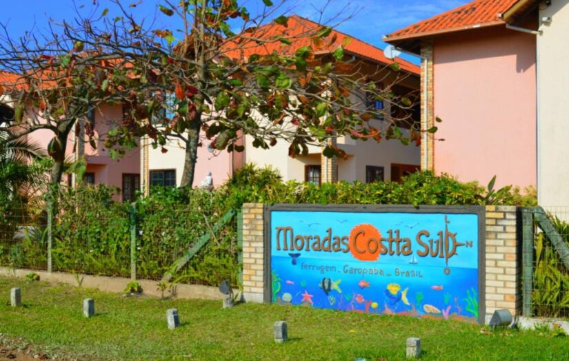 onde ficar em Garopaba na Praia do Ferrugem Uma placa colorida para Moradas Costa Sul, uma opção de hotel na Praia do Ferrugem, em Santa Catarina, em frente a casas cor-de-rosa com telhados vermelhos.
