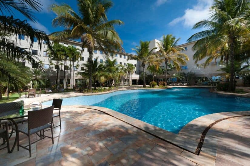 Piscina externa do hotel onde ficar em Goiânia cercada por palmeiras, com cadeiras e mesas no pátio de pedra sob um céu azul brilhante.