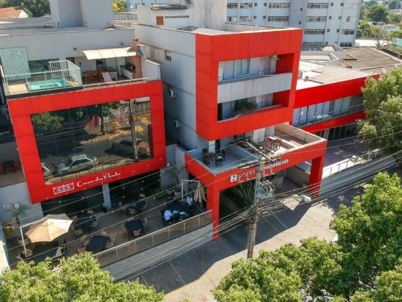 Moderno edifício vermelho e cinza de hotel com assentos ao ar livre, árvores e linhas de energia na frente.