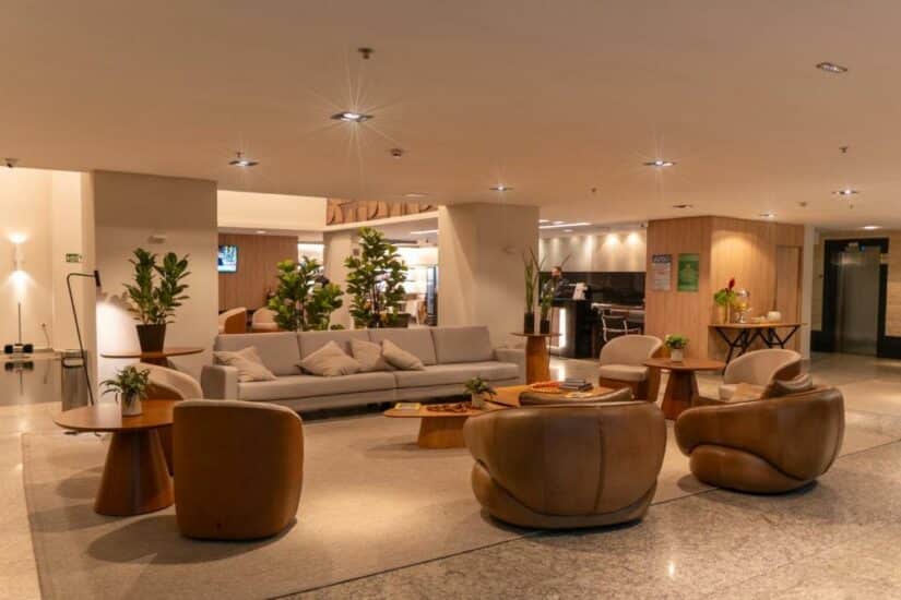 Saguão de hotel moderno onde ficar em Goiânia com cadeiras de couro bronzeado, um sofá cinza, vasos de plantas e iluminação aconchegante.