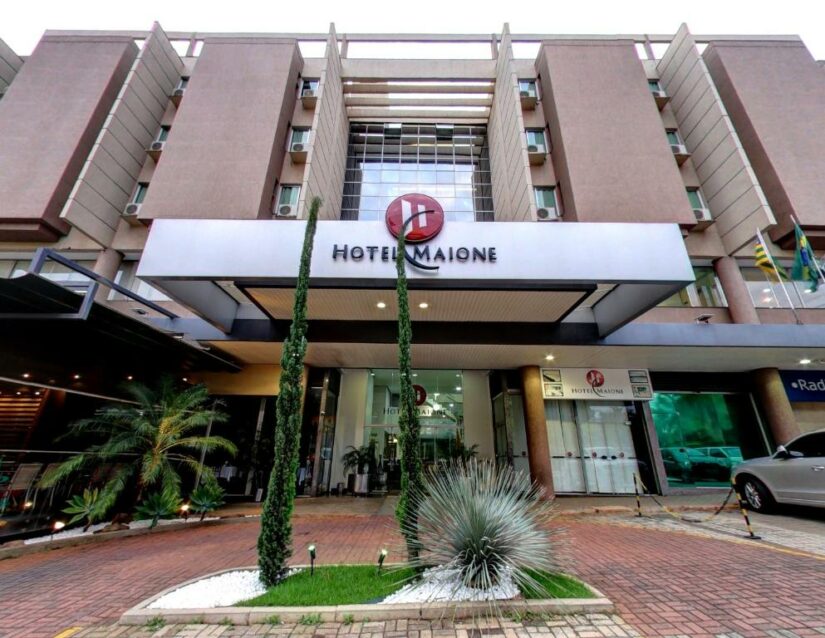 Entrada do Hotel Maione com plantas altas, portas de vidro e carros estacionados nas proximidades.
