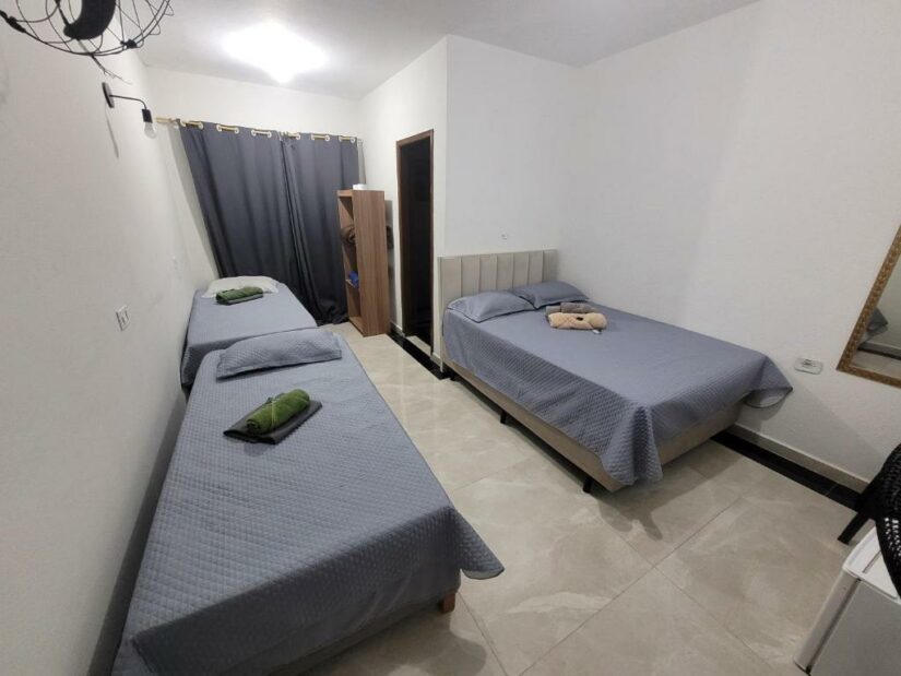 Um quarto simples com duas camas de solteiro e uma cama de casal onde ficar em Guararema, todas com roupa de cama cinza e toalhas por cima.