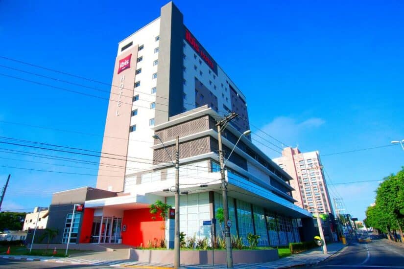 Edifício alto e moderno de hotel onde ficar em Guararema com detalhes em vermelho em um dia ensolarado, situado ao longo de uma rua da cidade com céu azul claro.