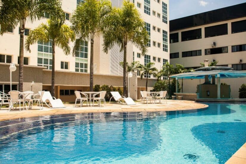 Piscina externa curva com espreguiçadeiras, mesas e palmeiras ao lado de um moderno edifício de hotel onde ficar em Guararema em um dia ensolarado.