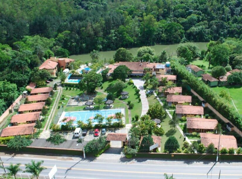 Vista aérea de um resort com edifícios de telhado vermelho, quadra de tênis, estacionamento, árvores e um rio ao fundo.