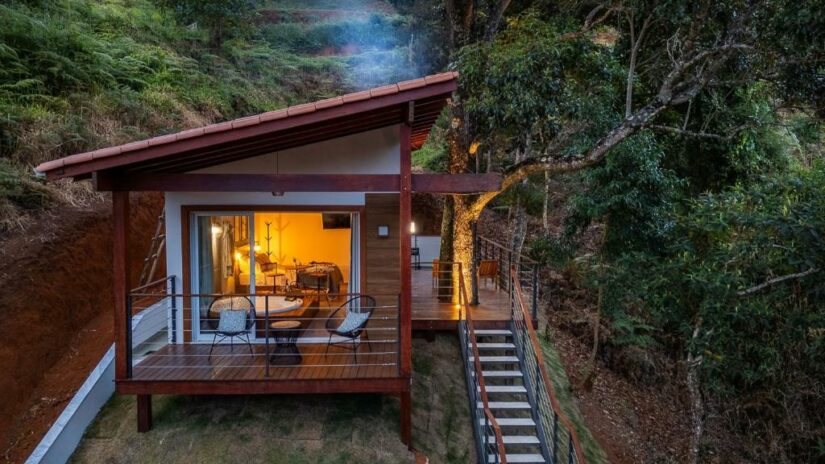 Uma pequena cabana moderna com interior iluminado, deck de madeira e escadas, cercada por árvores e vegetação ao anoitecer.