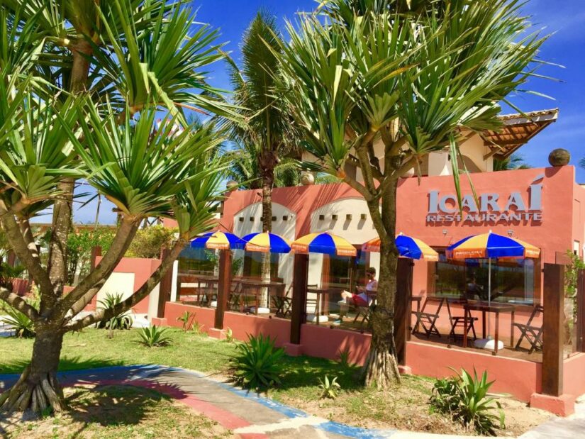 Restaurante com palmeiras onde ficar em Ilha Comprida, guarda-chuvas azuis-amarelos e assentos ao ar livre em um dia ensolarado.
