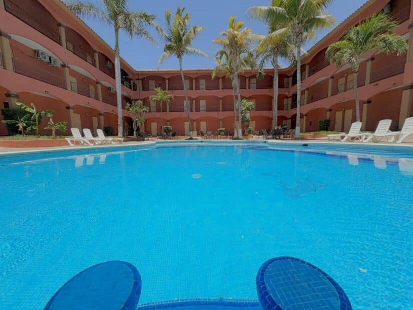 Uma piscina em um pátio ensolarado cercada por palmeiras, espreguiçadeiras e um prédio vermelho do hotel onde ficar em Los Cabos.