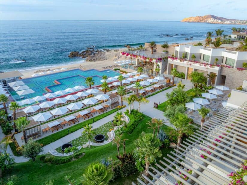 Vista aérea de um resort à beira-mar com piscina, espreguiçadeiras, guarda-sóis e paisagismo exuberante à beira-mar.