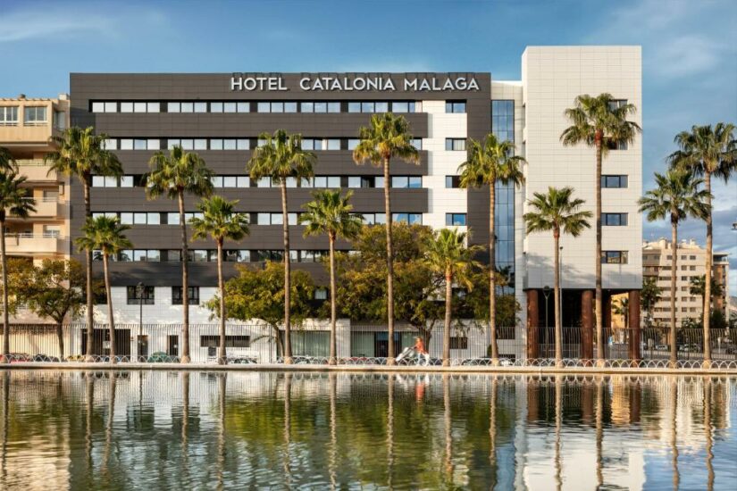 Um moderno edifício de hotel, o Hotel Catalonia Malaga, com palmeiras refletidas em uma piscina em frente.