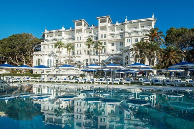 Luxuoso hotel branco onde ficar em Málaga com palmeiras, guarda-sóis azuis e uma piscina transparente que reflete o prédio e o céu.