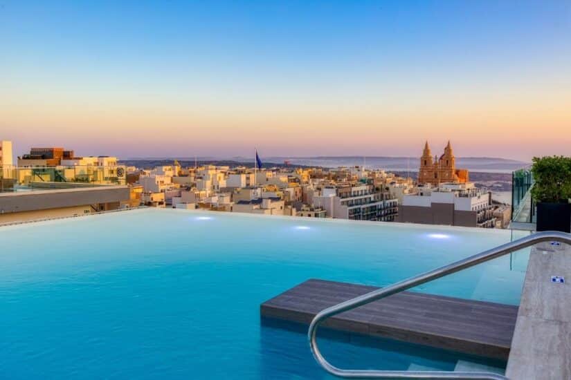 Piscina infinita na cobertura com vista para a paisagem urbana e a igreja distante ao pôr do sol.