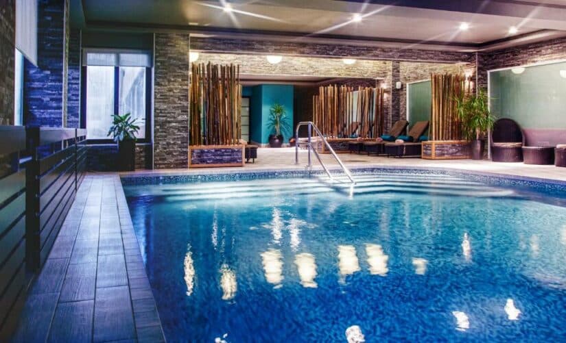 Piscina interna com espreguiçadeiras, plantas e divisórias de bambu em um ambiente de spa moderno e relaxante.