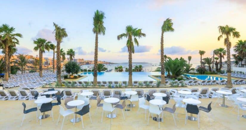 Área de estar externa com mesas brancas onde ficar em Malta, palmeiras e piscinas com vista para o mar ao pôr do sol.