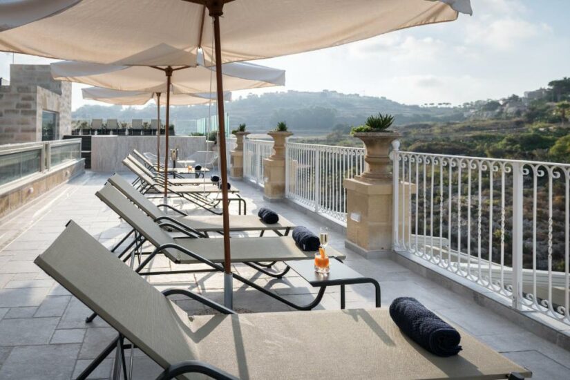 Espreguiçadeiras com toalhas e bebidas sob guarda-sóis em um terraço ensolarado com vista para um vale panorâmico.