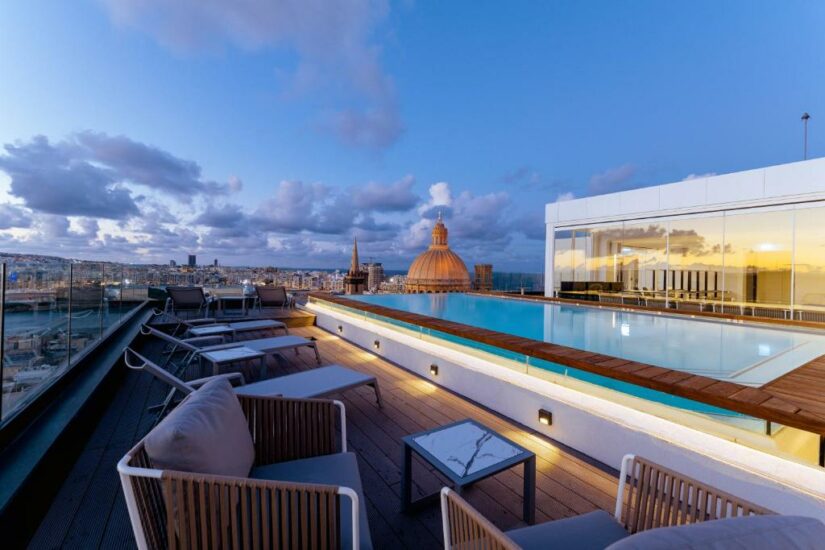 Piscina na cobertura com espreguiçadeiras onde ficar em Malta, paisagem urbana e edifício abobadado ao pôr do sol sob um céu parcialmente nublado.