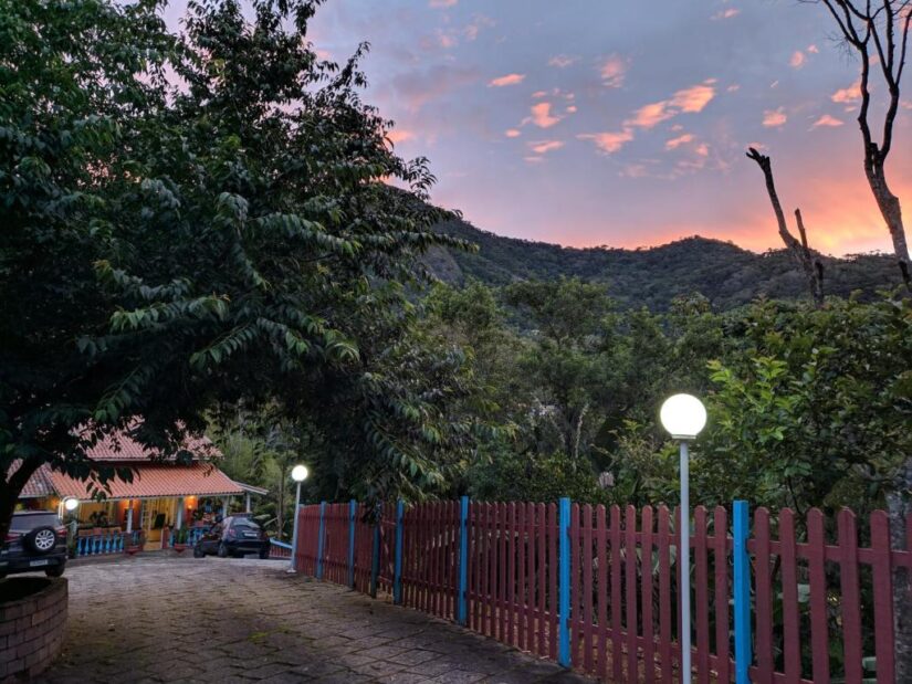 onde ficar em Nova Friburgo em Lumiar Uma trilha pavimentada com uma pousada em Lumiar no RJ vermelha, árvores, carros estacionados e colinas ao pôr do sol.