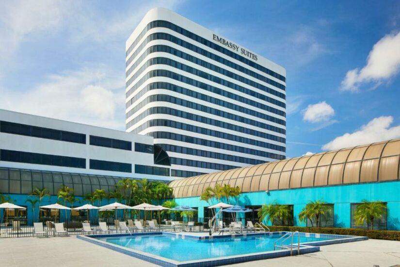 Piscina externa do hotel onde ficar em Palm Beach com espreguiçadeiras, guarda-sóis, palmeiras e um prédio alto do Embassy Suites ao fundo.