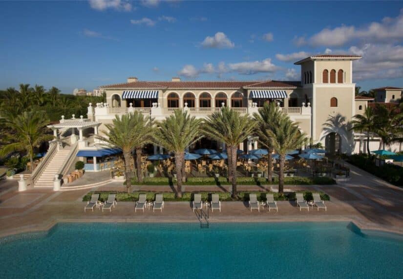 Edifício em estilo mediterrâneo onde ficar em Palm Beach com palmeiras, mesas ao ar livre e uma piscina em primeiro plano sob o céu azul.