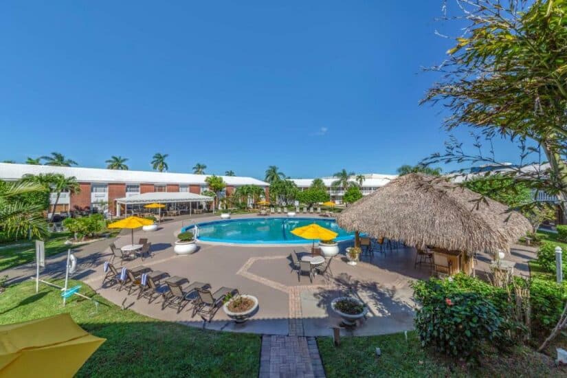 Área da piscina do resort com espreguiçadeiras, guarda-sóis amarelos, palmeiras e uma cabana com telhado de palha sob um céu azul claro.