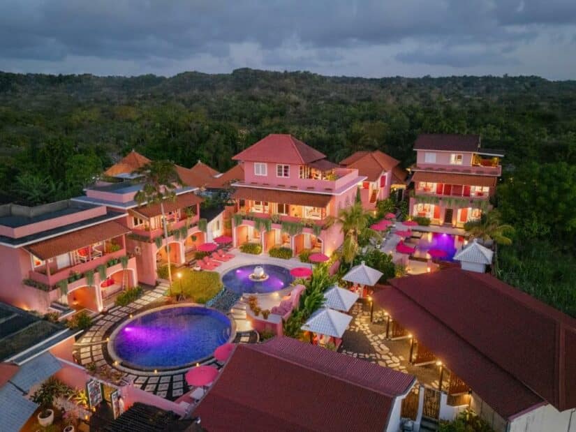 Vista aérea de um resort colorido com edifícios cor-de-rosa, piscinas e assentos ao ar livre, cercado por vegetação ao anoitecer.